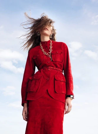 Model wearing a red belted jacket and skirt against a cloudy blue sky with hair blowing in the wind