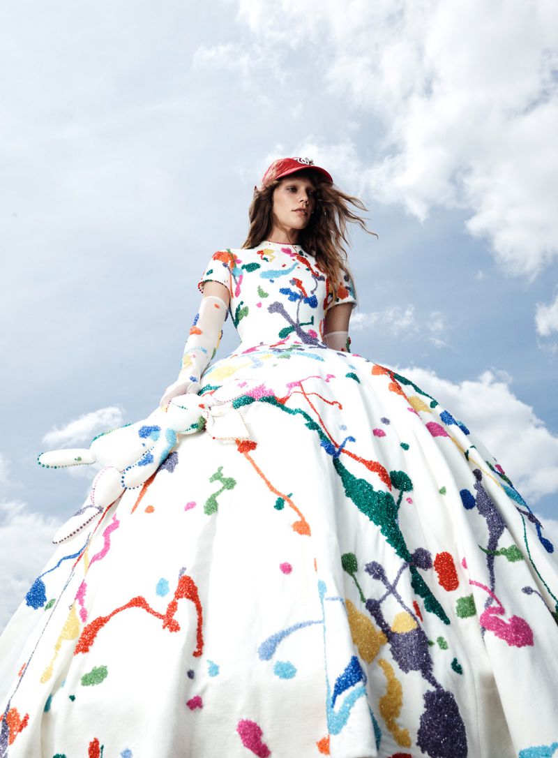 Model in a voluminous white paint-splattered gown and red cap photographed from below against a cloudy sky.
