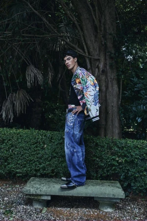 Model in floral print bomber jacket and distressed blue denim standing on stone bench outdoors against trees