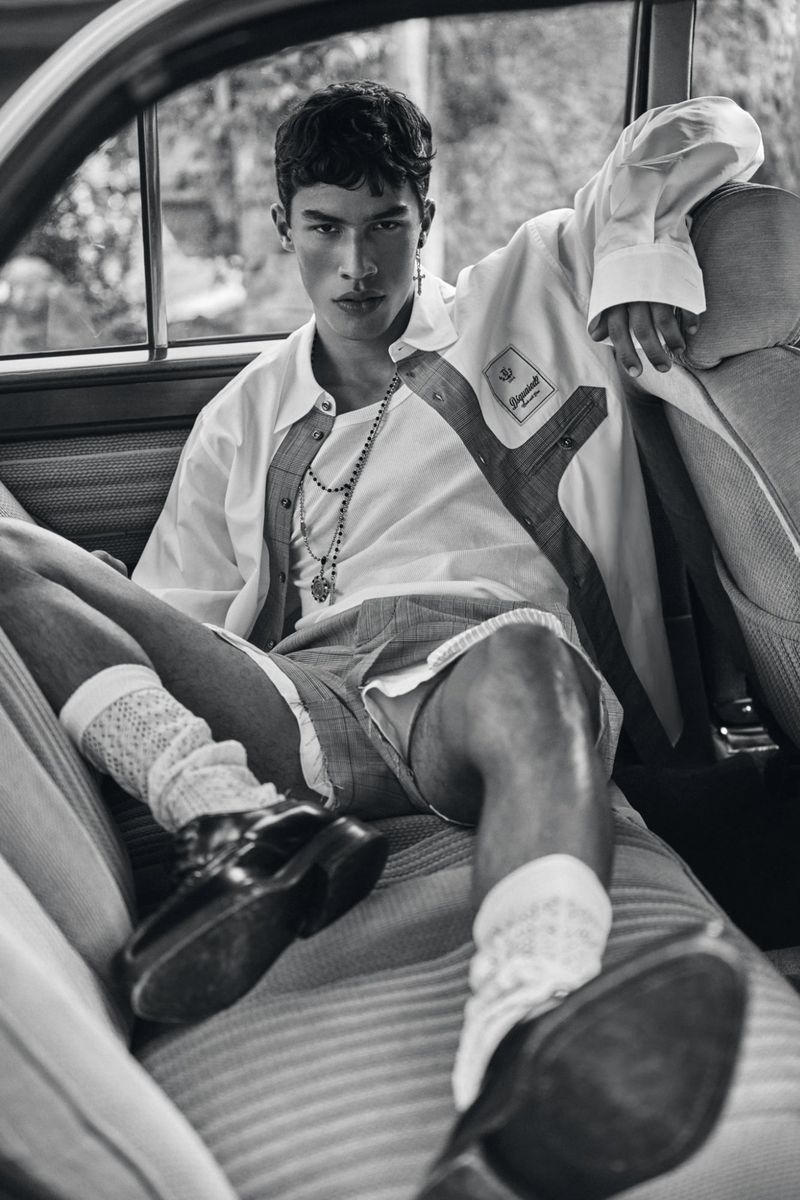 Model seated inside a car wearing an unbuttoned shirt, shorts, layered necklaces, patterned socks, and shoes.