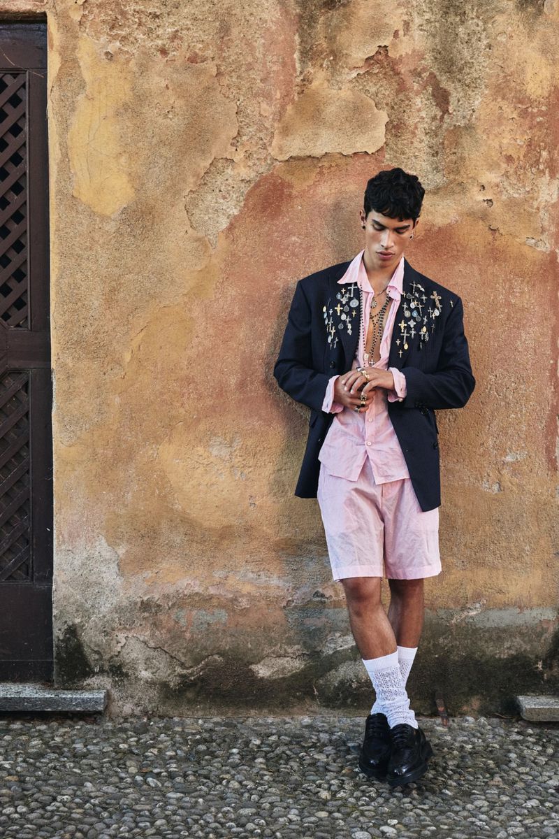 Male model in pink shirt and dark blazer leaning against textured stone wall outdoors for Dsquared2