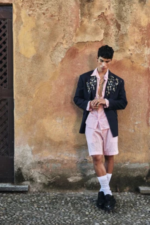 Male model in pink shirt and dark blazer leaning against textured stone wall outdoors for Dsquared2
