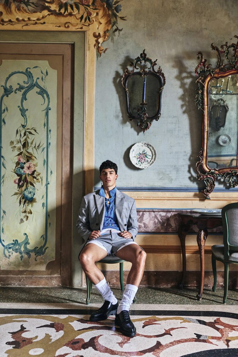 A seated male model in a grey suit jacket and shorts posing in an ornate, frescoed interior room.