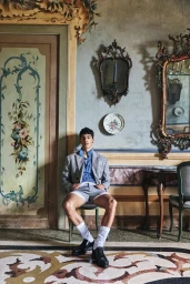 A seated male model in a grey suit jacket and shorts posing in an ornate, frescoed interior room.