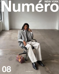 Model seated on an office chair wearing a grey blazer, white shirt, and tie, with a monogram handbag placed beside them on a concrete floor.