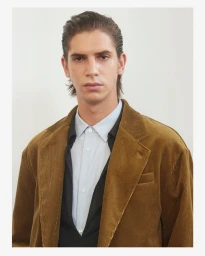 Portrait of a man wearing a brown corduroy blazer over a collared shirt against a white wall.