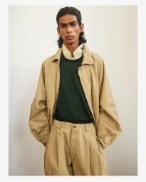 Model wearing a tan jacket, green shirt, and matching trousers against a plain light wall.