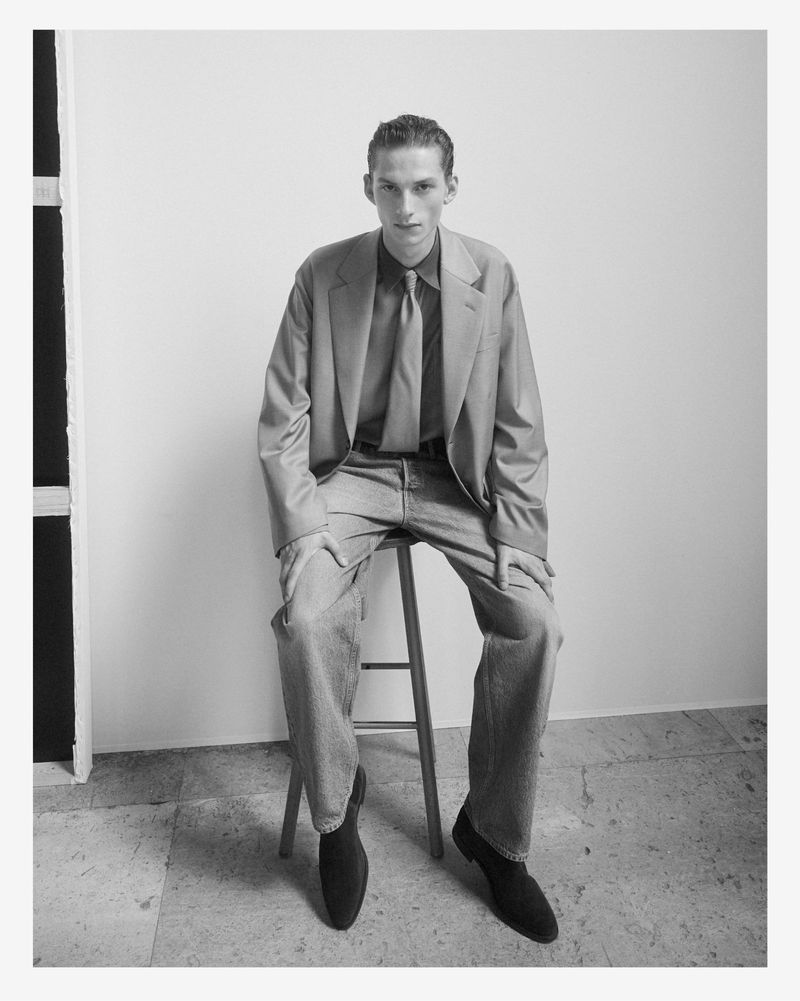 Monochrome portrait of a male model seated on a stool wearing a tailored suit jacket, tie, and trousers against a plain wall.