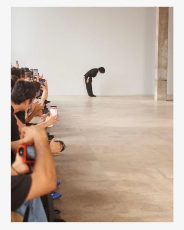 A designer bowing at the end of a runway show before an audience of photographers seated along the side.