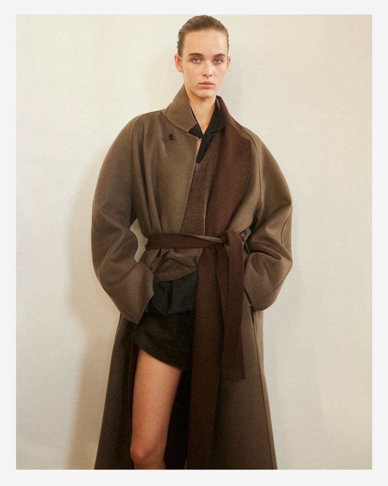 Model wearing a structured brown belted coat over dark layers against a pale seamless studio background