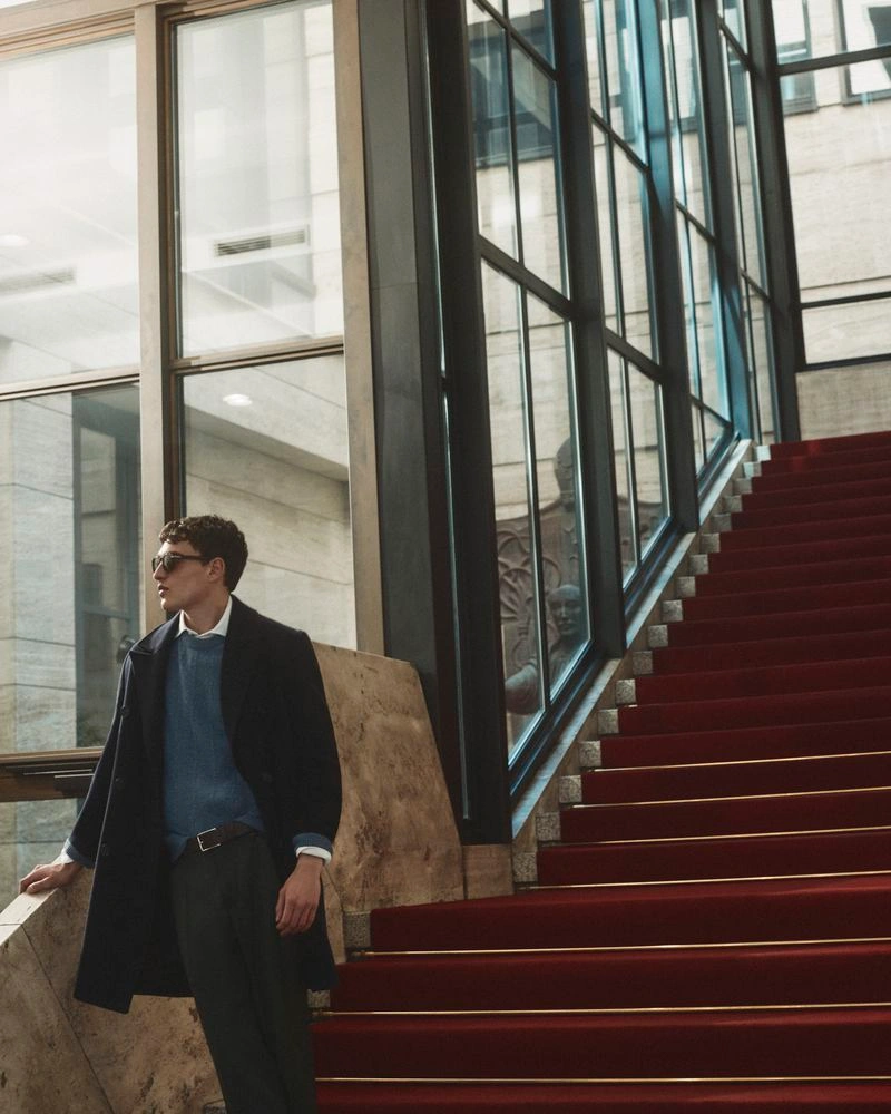 Man standing on a grand red staircase next to tall glass windows wearing a dark coat and blue sweater for Manuel Ritz FW26.