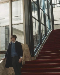 Man standing on a grand red staircase next to tall glass windows wearing a dark coat and blue sweater for Manuel Ritz FW26.