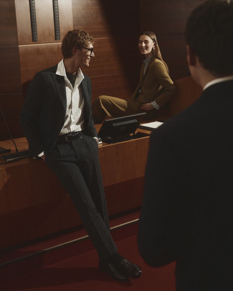 Man leaning against a wood desk in a suit with a woman and another man in an interior setting for Manuel Ritz FW26.