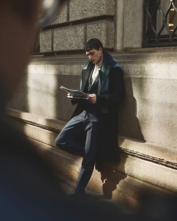 Man wearing a dark coat and suit reading a newspaper while leaning against a stone building exterior