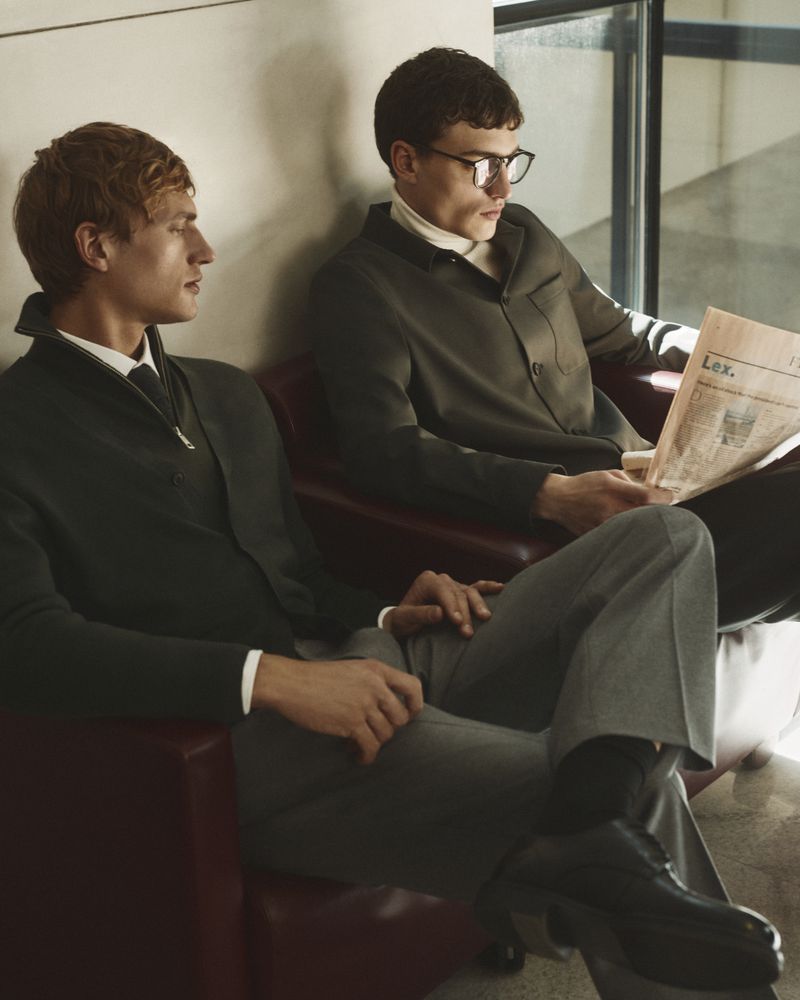Two male models seated in red armchairs, one reading a newspaper for Manuel Ritz FW26 campaign by Gautier Pellegrin.