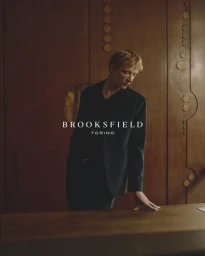 Blonde male model leaning on a wooden table in a dark blazer against a paneled wood wall for Brooksfield FW26.