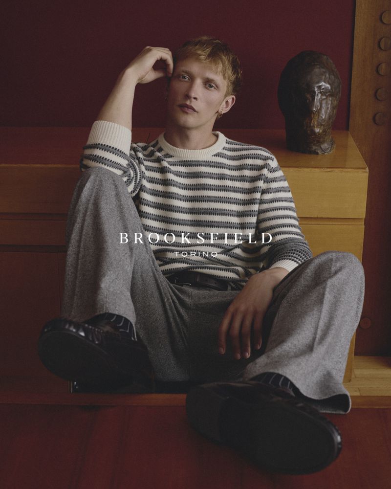 Model seated on a wooden cabinet wearing a striped crewneck sweater and grey trousers for Brooksfield FW26.