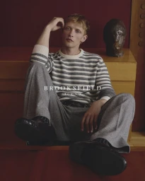 Model seated on a wooden cabinet wearing a striped crewneck sweater and grey trousers for Brooksfield FW26.