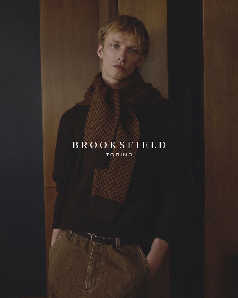 A male model wearing a brown knit scarf and dark sweater posing against wood paneling for Brooksfield FW26.