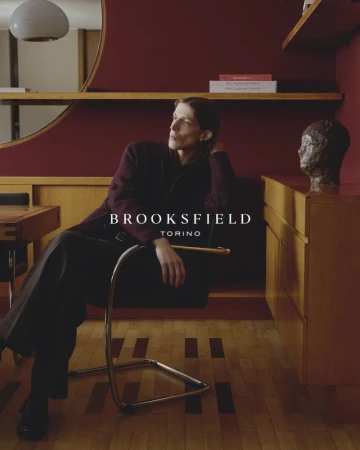 Model seated in a modern cantilever chair inside a warmly lit room with deep red walls and wood paneling.