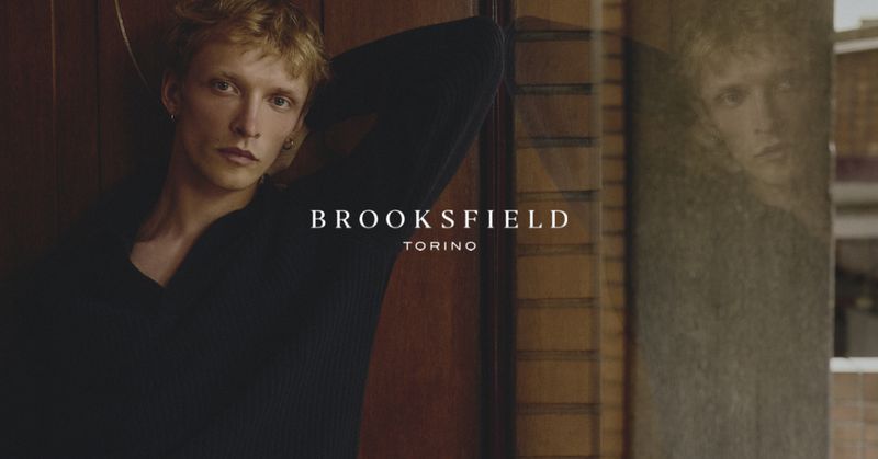 Blonde male model wearing a dark ribbed sweater leaning against a wall indoors for Brooksfield