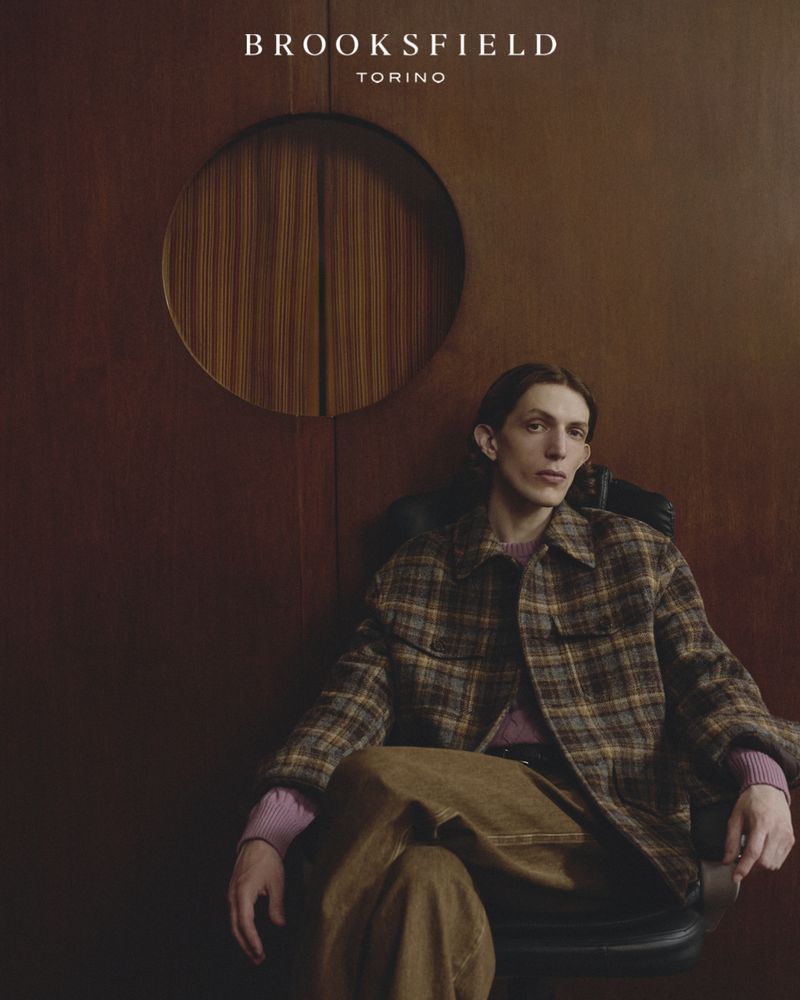 A man wearing a plaid jacket and brown trousers seated indoors against a dark wood wall with a circular cutout panel.