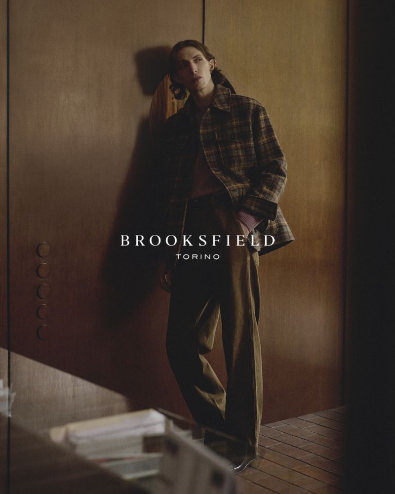 Model leaning against a dark wood wall wearing a plaid jacket and brown trousers for Brooksfield FW26.