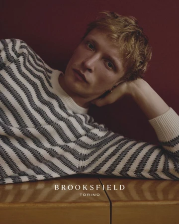 Blonde male model wearing a striped knit sweater resting his head on his hand against a deep red wall