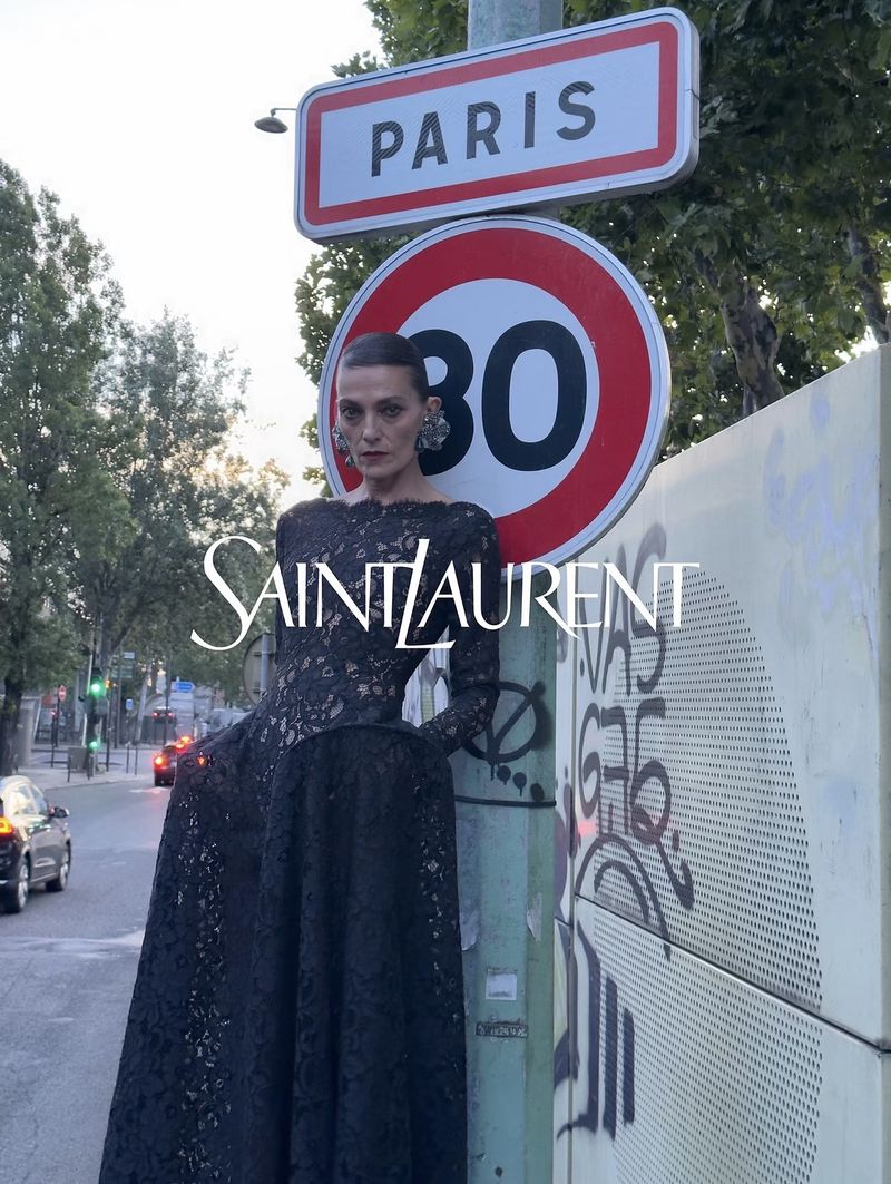 Model in a black lace dress standing on a Paris street leaning against a traffic sign for Saint Laurent Winter 2026 campaign.