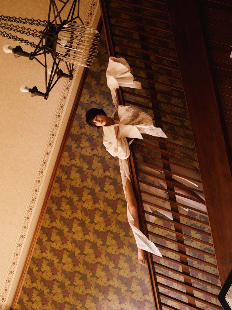 Model in flowing white dress leaning against a dark wooden staircase railing under an ornate chandelier.