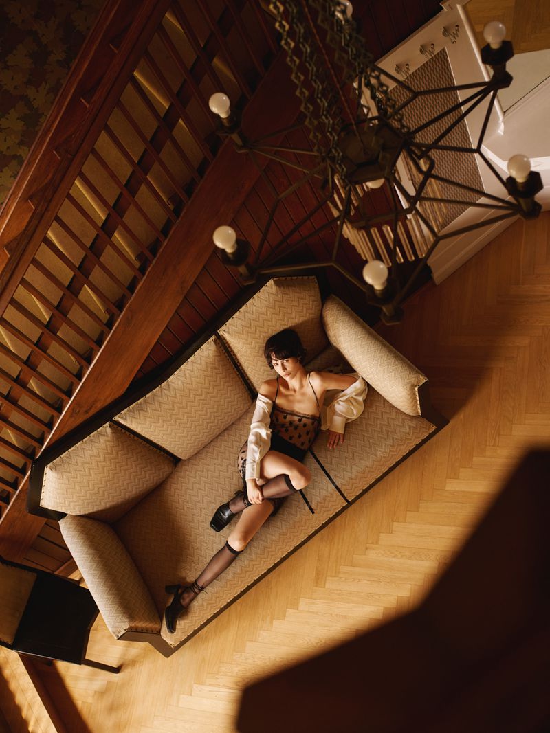 High angle shot of a woman lounging on a patterned sofa near a wooden staircase and large chandelier