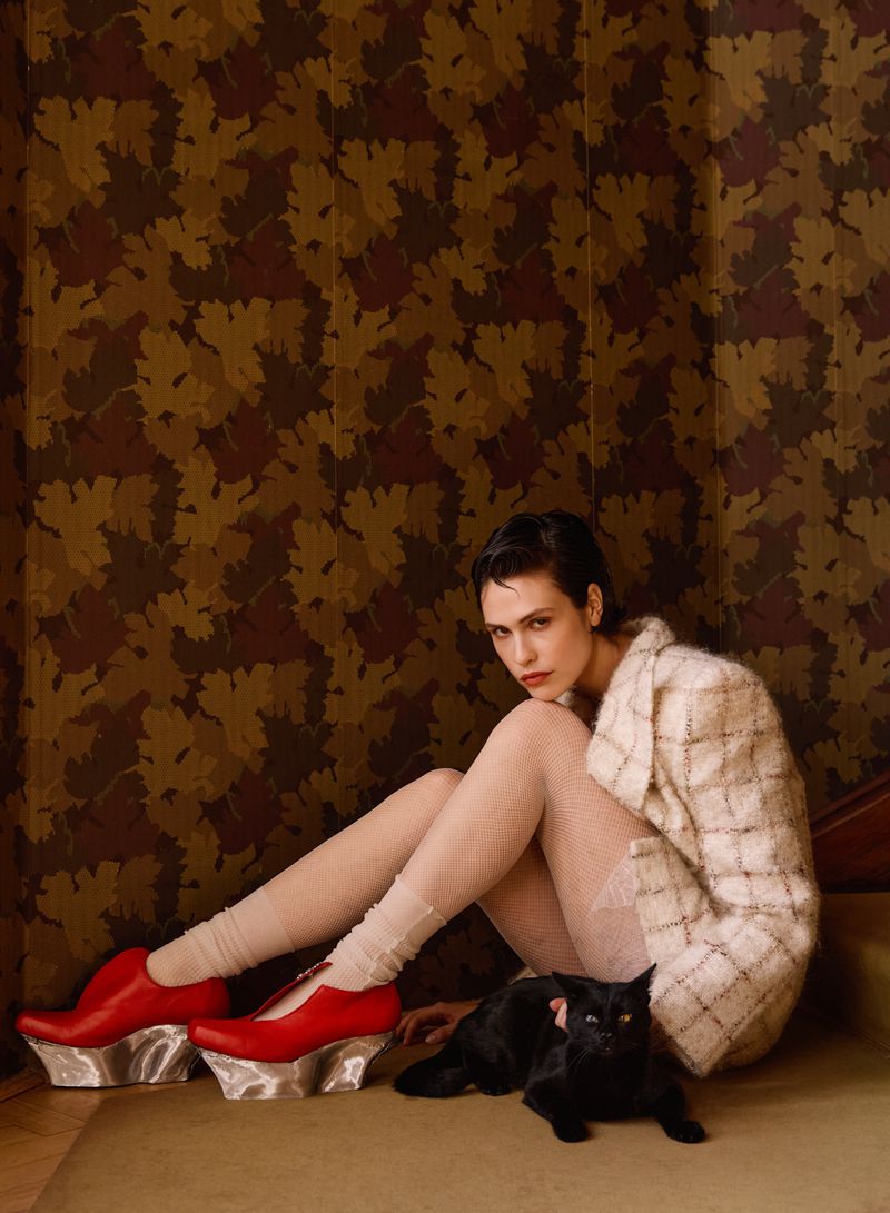 Model wearing a plaid jacket and bright red platform shoes seated beside a black cat against patterned wallpaper.