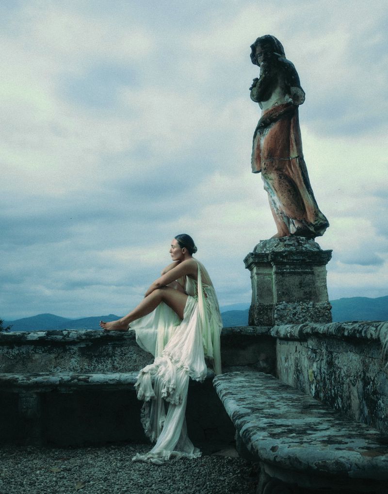 Woman in flowing white dress sits on stone wall with statue backdrop, enjoying the view of rolling hills and cloudy sky.