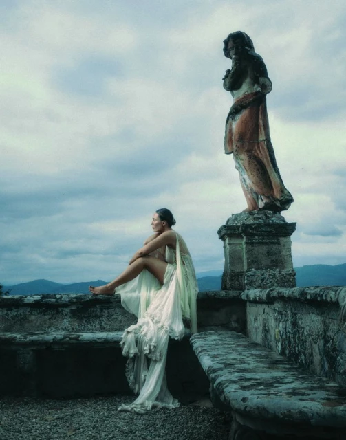 Woman in flowing white dress sits on stone wall with statue backdrop, enjoying the view of rolling hills and cloudy sky.