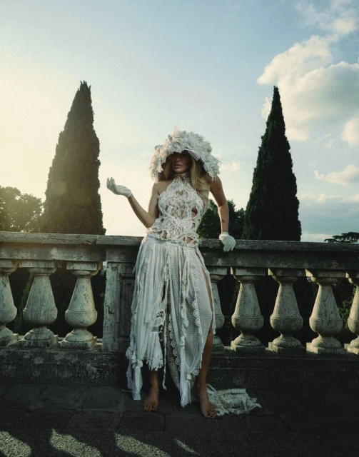 A blonde woman wears a white couture dress and hat, posing on a stone balcony with cypress trees under a blue sky with light clouds.