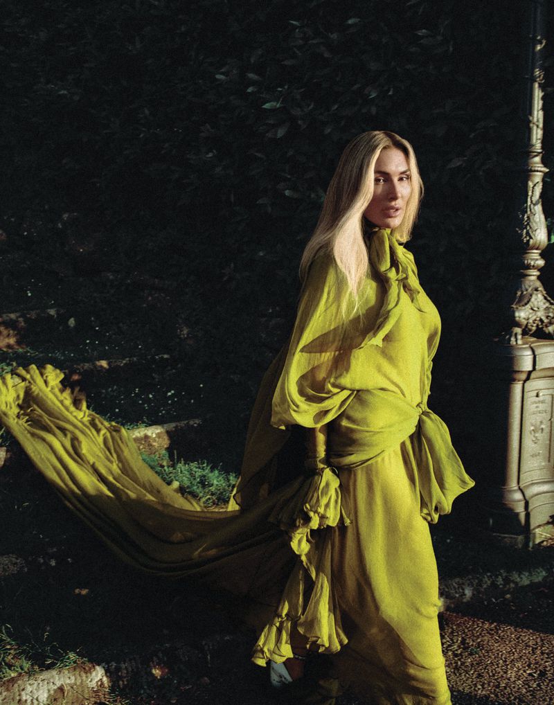A fair-skinned woman in a long chartreuse gown stands on stone steps in a garden, with dappled lighting and dark leafy background.