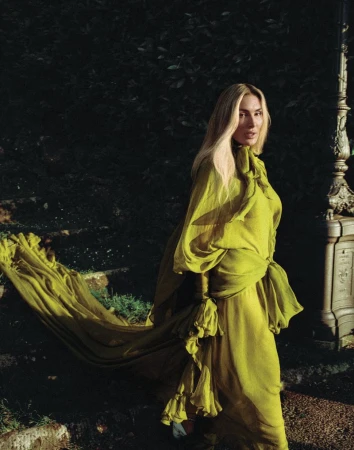 A fair-skinned woman in a long chartreuse gown stands on stone steps in a garden, with dappled lighting and dark leafy background.