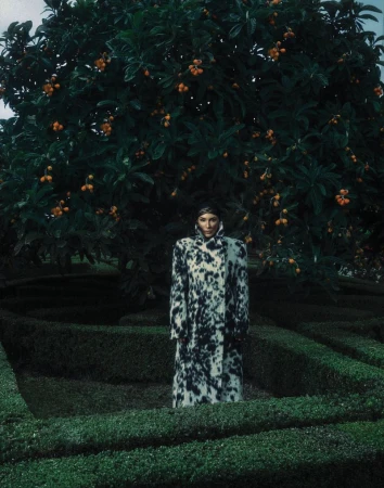 Woman in patterned coat stands in a manicured garden under a large tree with orange fruit. Fashion editorial photography.