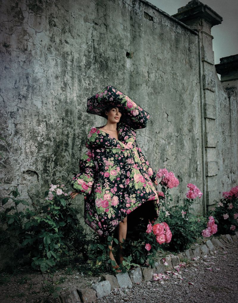 A woman in a floral print coat and large hat stands in a garden with pink roses against a weathered grey wall.