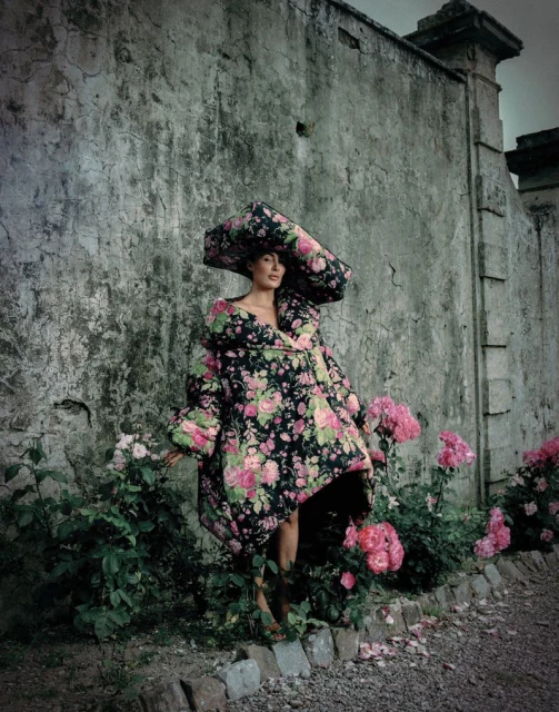 A woman in a floral print coat and large hat stands in a garden with pink roses against a weathered grey wall.