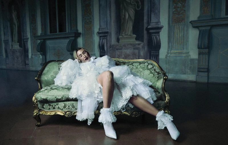 An adult woman rests on a vintage green velvet sofa wearing a frilly white dress and boots in an ornate room with wall murals.