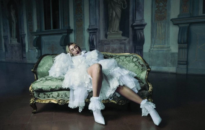 An adult woman rests on a vintage green velvet sofa wearing a frilly white dress and boots in an ornate room with wall murals.