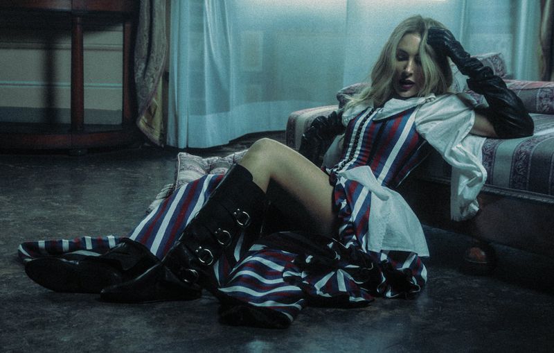 Blonde woman in a striped corset and black boots reclines on a striped chaise lounge in a dim room with a dark wood floor and white curtains.