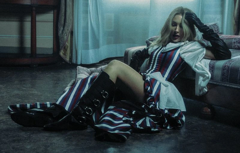 Blonde woman in a striped corset and black boots reclines on a striped chaise lounge in a dim room with a dark wood floor and white curtains.