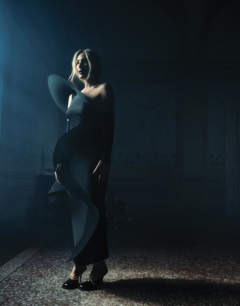 A woman in an elegant black dress with a large ruffle stands in a dark interior space, lit by a single spotlight.