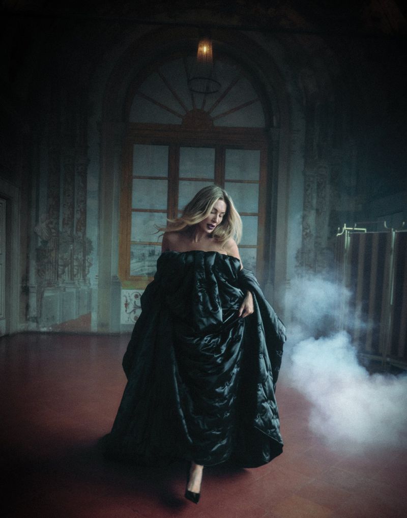 A blonde woman wearing a black puffy dress and high heels stands in a grand, empty room with a window and diffused lighting.