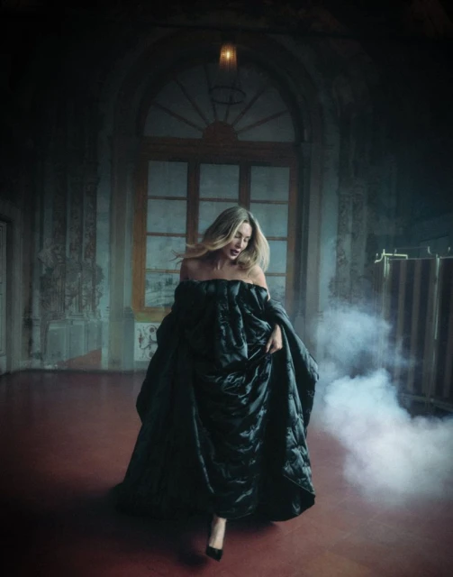 A blonde woman wearing a black puffy dress and high heels stands in a grand, empty room with a window and diffused lighting.