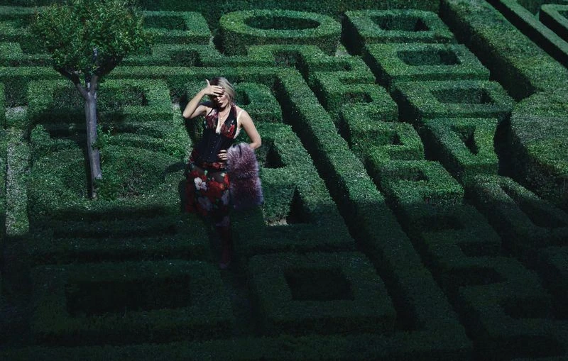 Woman in floral dress stands in the center of a lush green hedge maze with geometric shapes and shadows, creating a surreal scene.