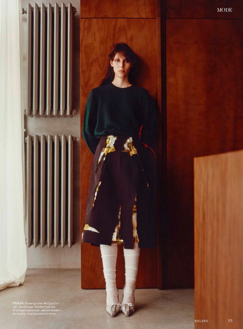 Model standing against wood wall in dark green sweater, patterned skirt, white socks, and heels beside radiators