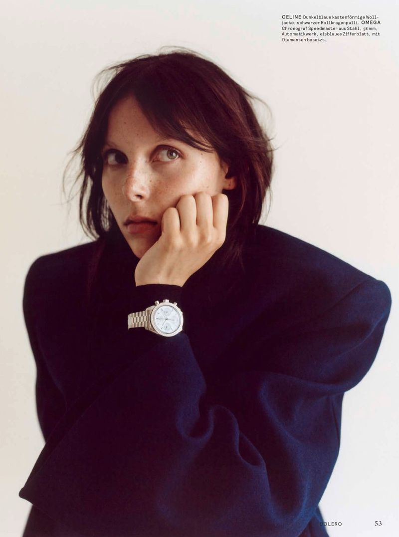 Close-up portrait of a woman with dark hair wearing a navy blue blazer and silver watch against a plain white background.
